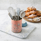MARIA ROMA Botanica small floral storage bin used as a baking tool holder with whisks, spatulas, and a sifter on a flour-dusted marble board with croissants in the background.